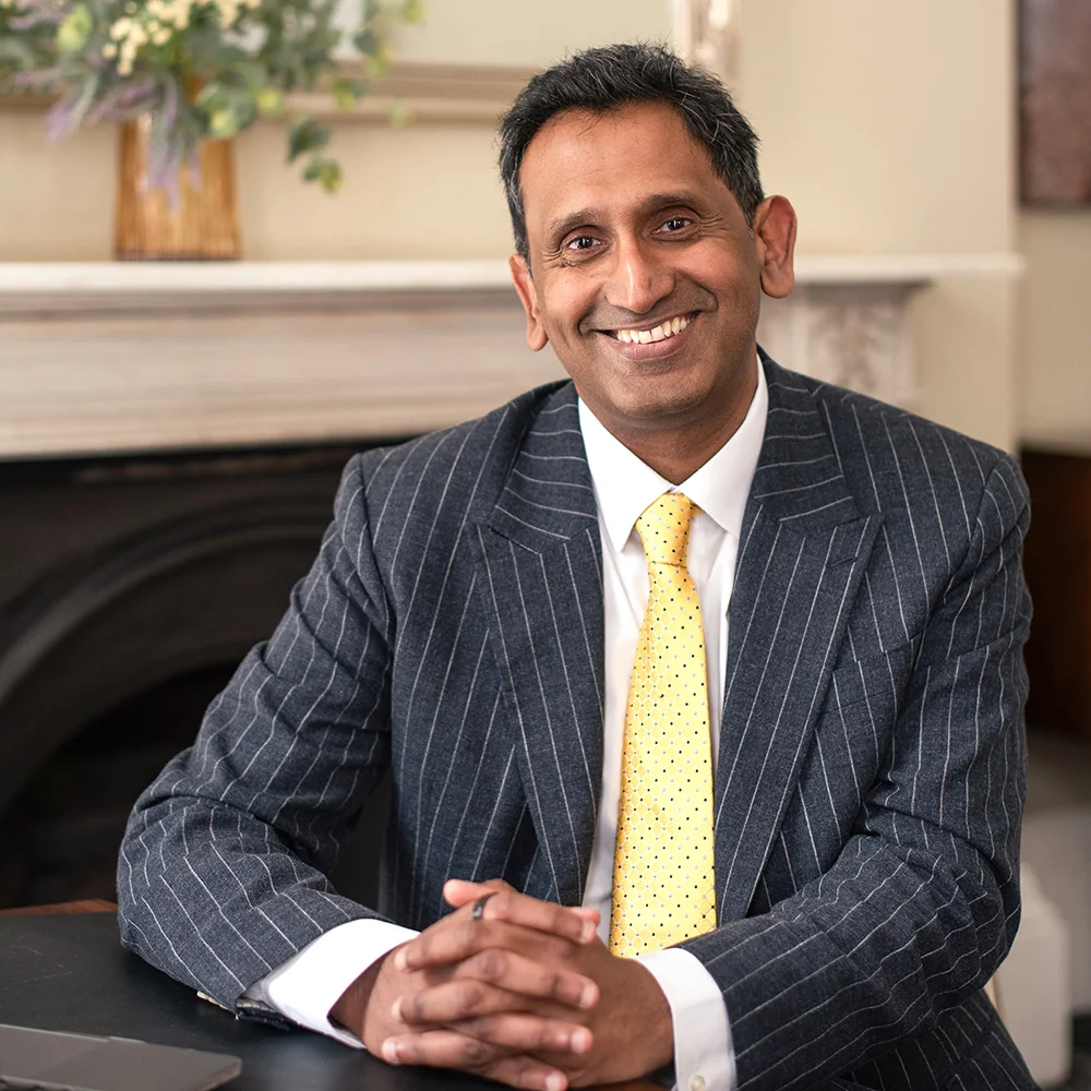 Professor Prasanna Sooriakumaran seated in his clinic, smiling with hands clasped